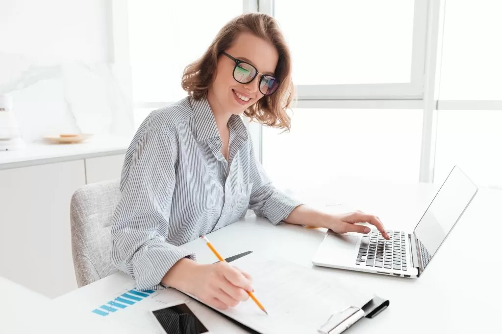 young-smiling-woman-in-glasses-and-striped-shirt-working-with-documents-and-computer-while-siting-at-table-in-light-kitchen.jpg